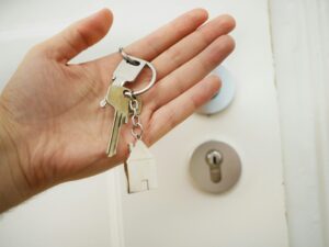 A person holds a set of house keys with a small house-shaped keychain in front of a white door with a modern lock, symbolizing home security and locksmith services.
