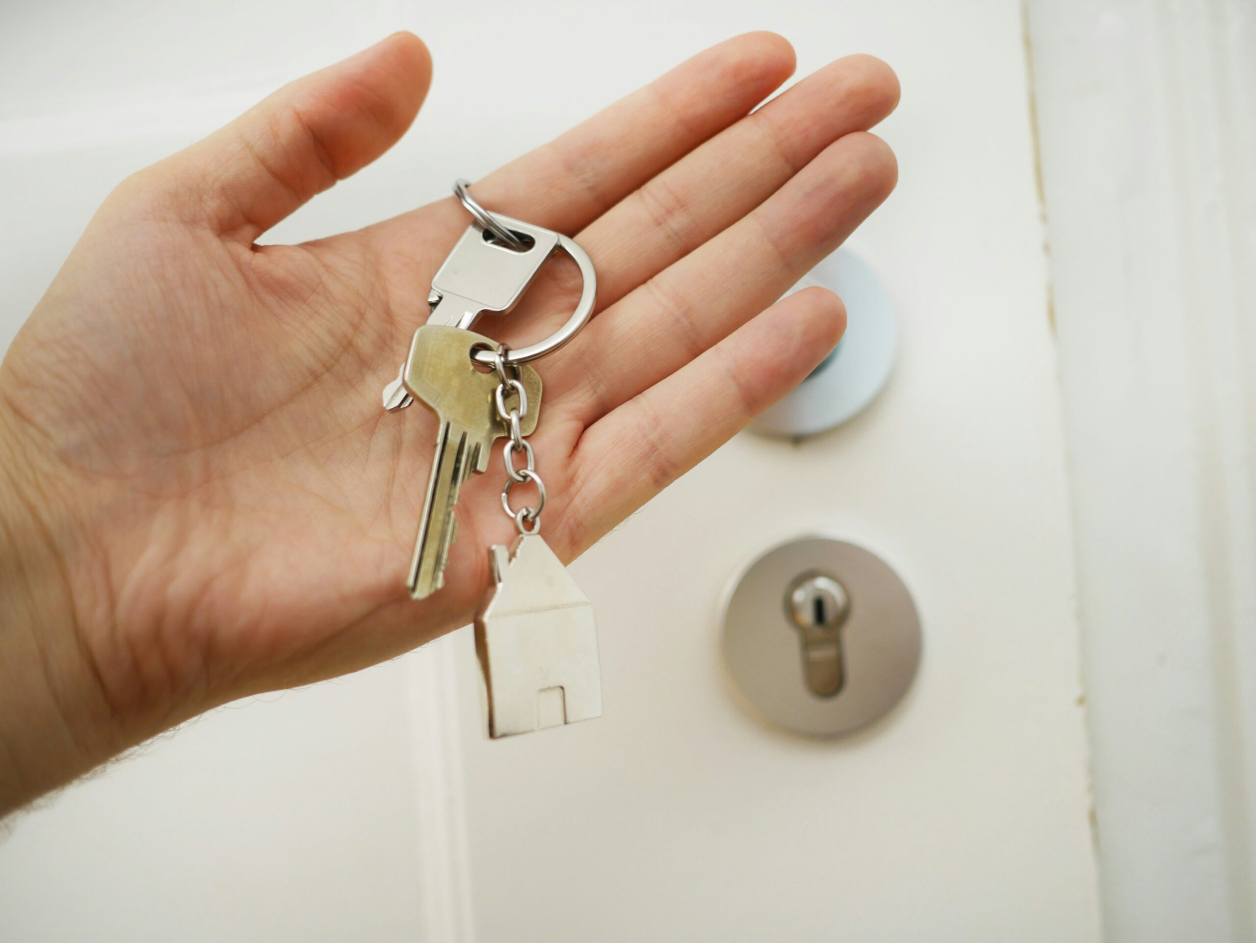 A person holds a set of house keys with a small house-shaped keychain in front of a white door with a modern lock, symbolizing home security and locksmith services.