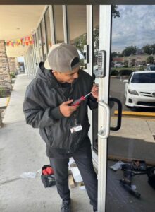 A professional locksmith in a black jacket and cap installs or repairs a keypad lock on a Residential glass door, using a screwdriver. Locksmith tools and equipment are scattered on the ground, highlighting the expertise and precision required for locksmith services.