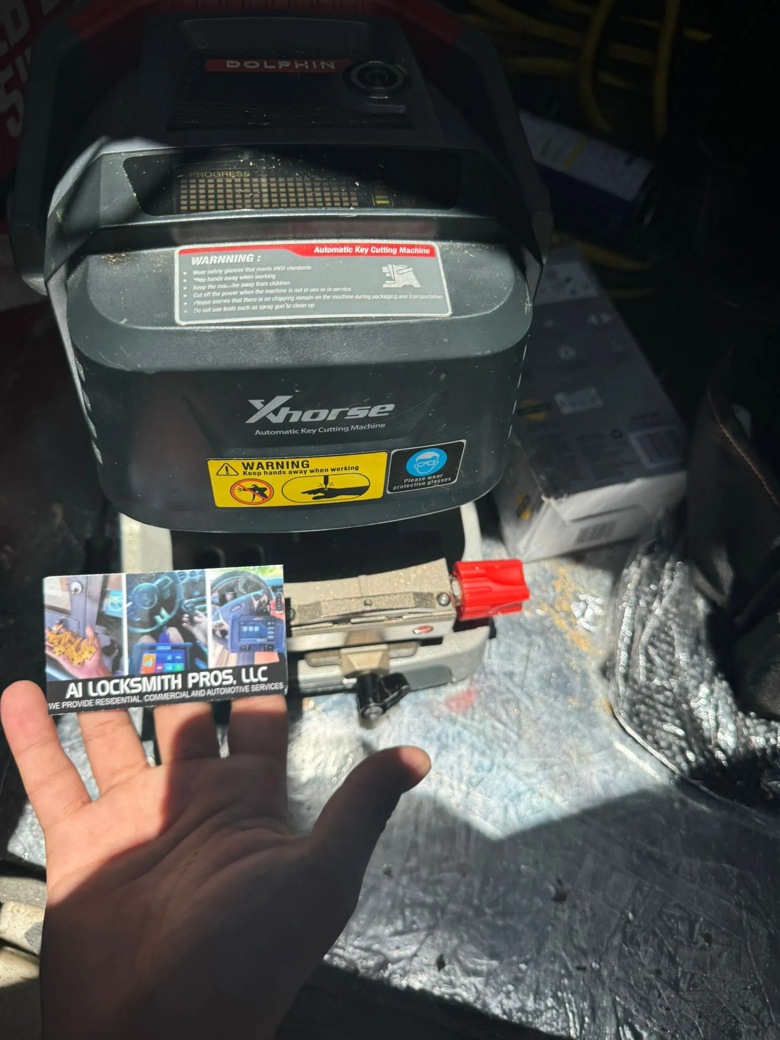 A1 Locksmith Service LLC technician holding a business card in front of a Xhorse Dolphin automatic key cutting machine used for professional key duplication and locksmith service LLC operations.