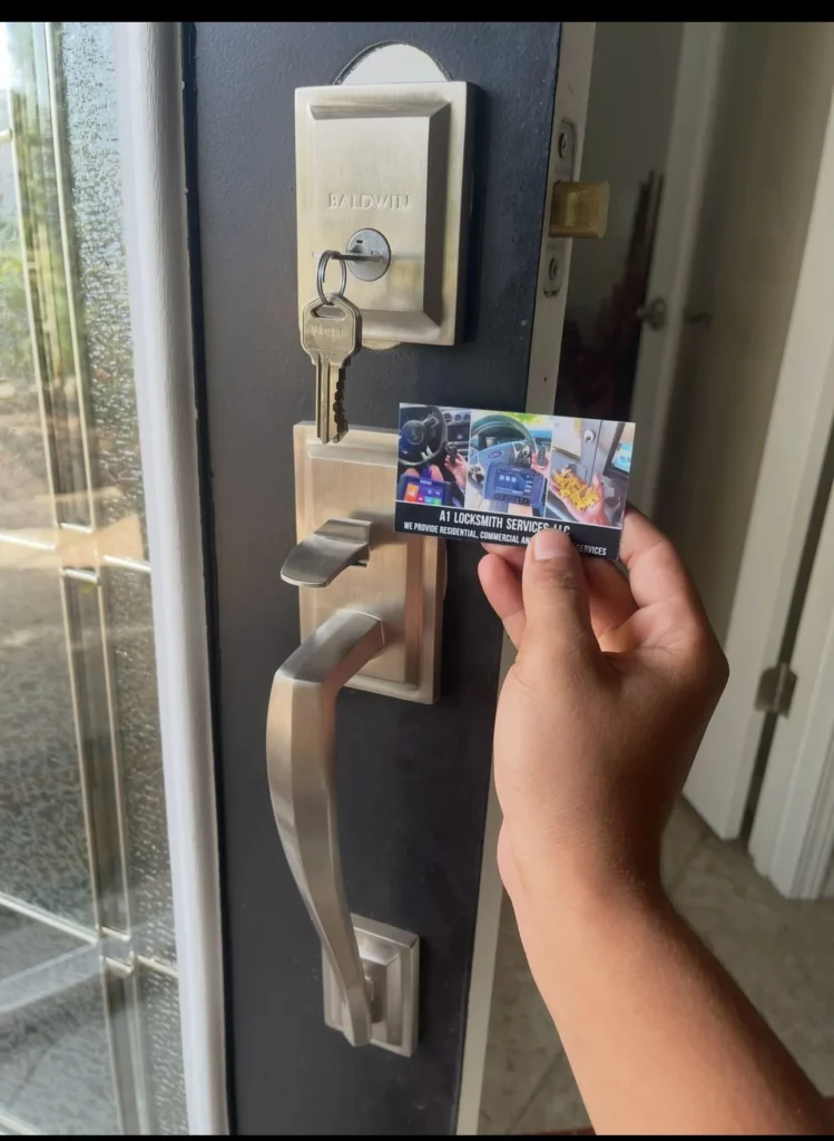 A hand holding an A1 Locksmith Services business card in front of a door lock with a Baldwin brand key inserted into the lock. The door has a modern brushed nickel handle and lock set.