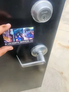 A hand holding an A1 Locksmith Services business card in front of a door with a Hager brand key lock and a modern silver lever handle. The background shows a concrete surface